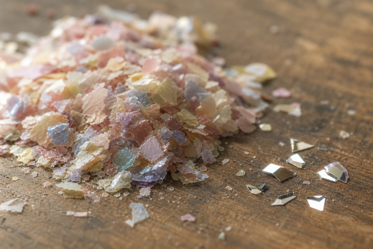 Détail d'un petit tas de bio-paillettes en cellulose plus mates, à côté de quelques particules synthétiques en PET brillantes, sur une surface en bois.